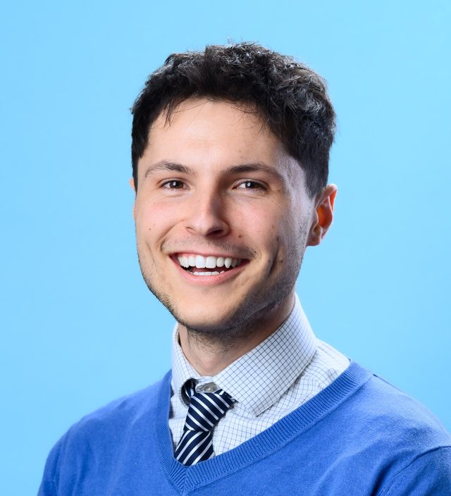 Aaron Agostini smiling in a blue sweater and striped tie against a light blue background